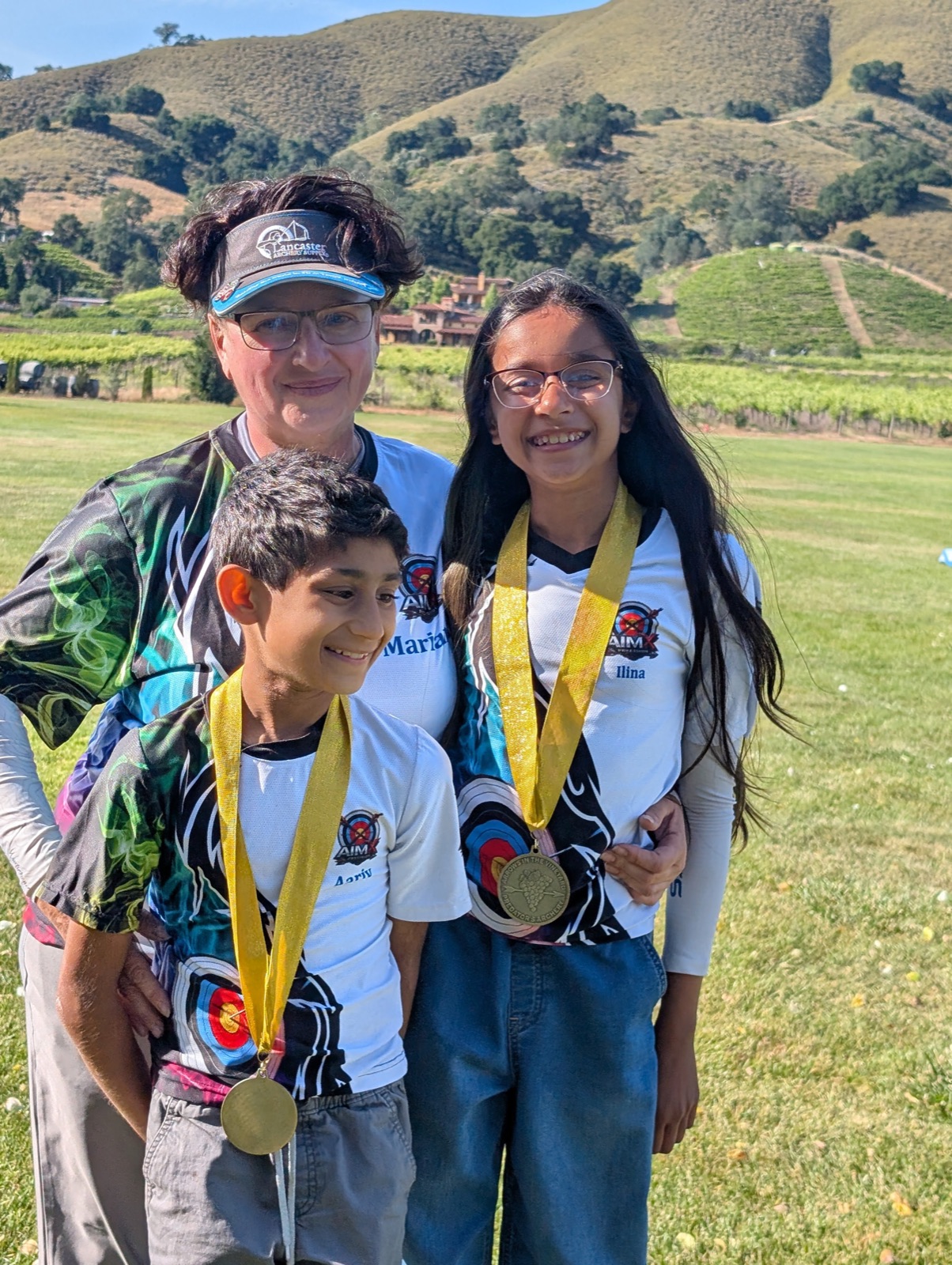 Coach Marianna with two AimX Precision students wearing their medals at an outdoor archery tournament